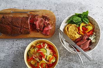 Un filet de bœuf balsamique sur une planche à découper en bois, à côté d’un bol de légumes et d’un autre de service composé de polenta, de légumes et de lanières de porc.