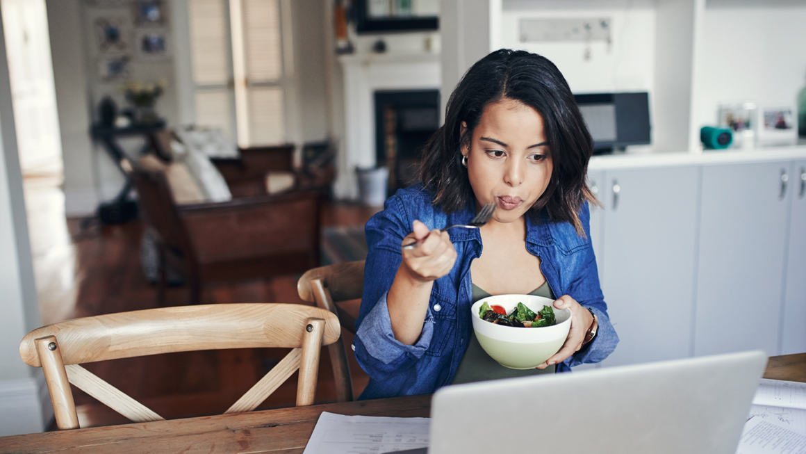 A young woman using a laptop and having a salad while working from home