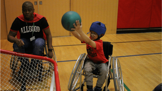 Un élève en fauteuil roulant soulève un ballon au-dessus de sa tête en visant un filet dans le gymnase d’une école. 