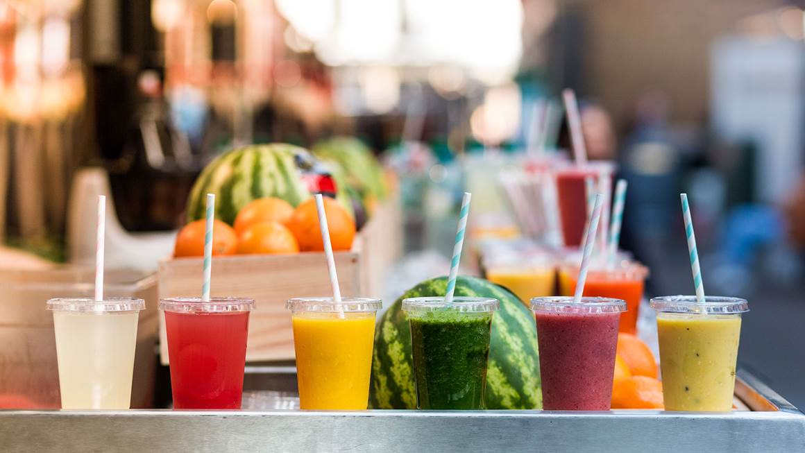 Six gobelets en plastique transparent remplis de jus de fruits colorés se trouvent devant divers fruits.