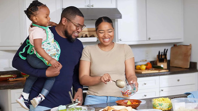 Une famille de trois est debout à côté d’un comptoir de cuisine; la mère prépare le repas et le père tient leur enfant d’un bras.