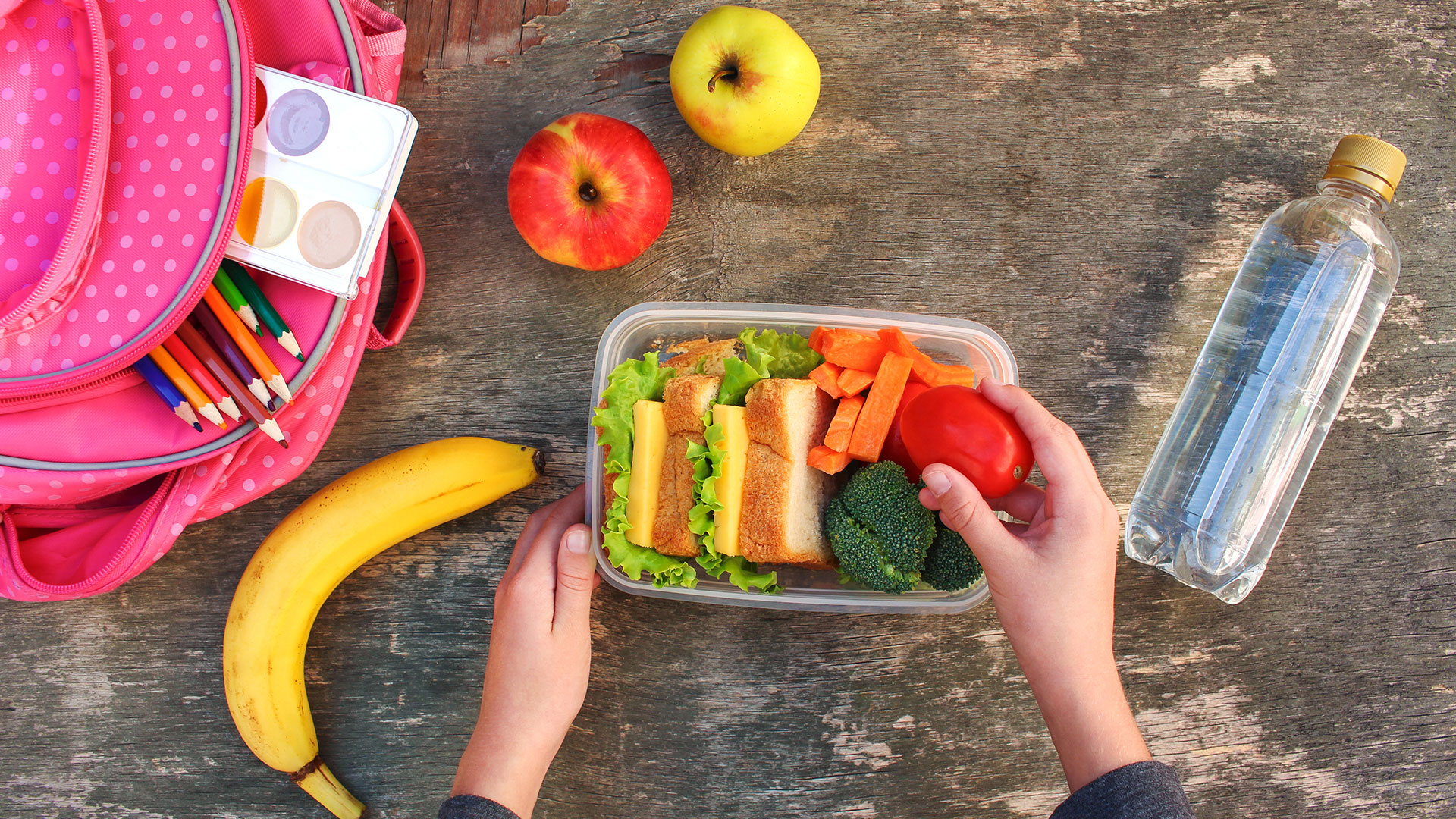Sandwichs, fruits et légumes dans un tupperware en plastique avec un sac à dos et une bouteille d'eau à côté sur un fond en bois.