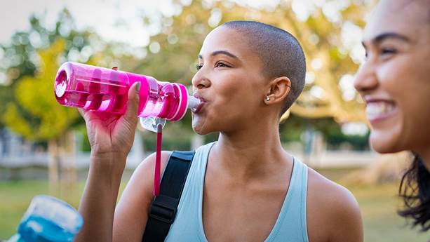 Jeune femme buvant de l’eau d’une bouteille réutilisable rose après avoir fait de l’exercice avec des amis dans un parc.