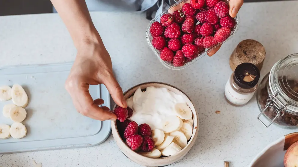 Une personne dépose des fraises dans un bol avec du yogourt et des bananes.