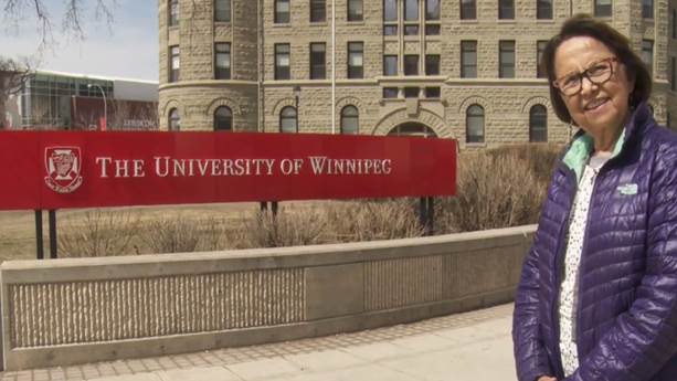 Esther Sanderson sourit, habillée d’une veste violette, à côté d’un panneau de l’Université de Winnipeg. 
