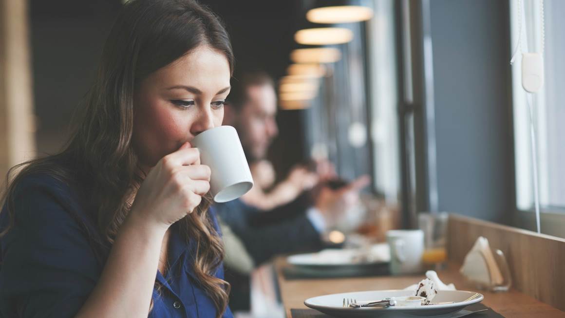 Une femme assise dans un restaurant sirote un café dans une tasse blanche.