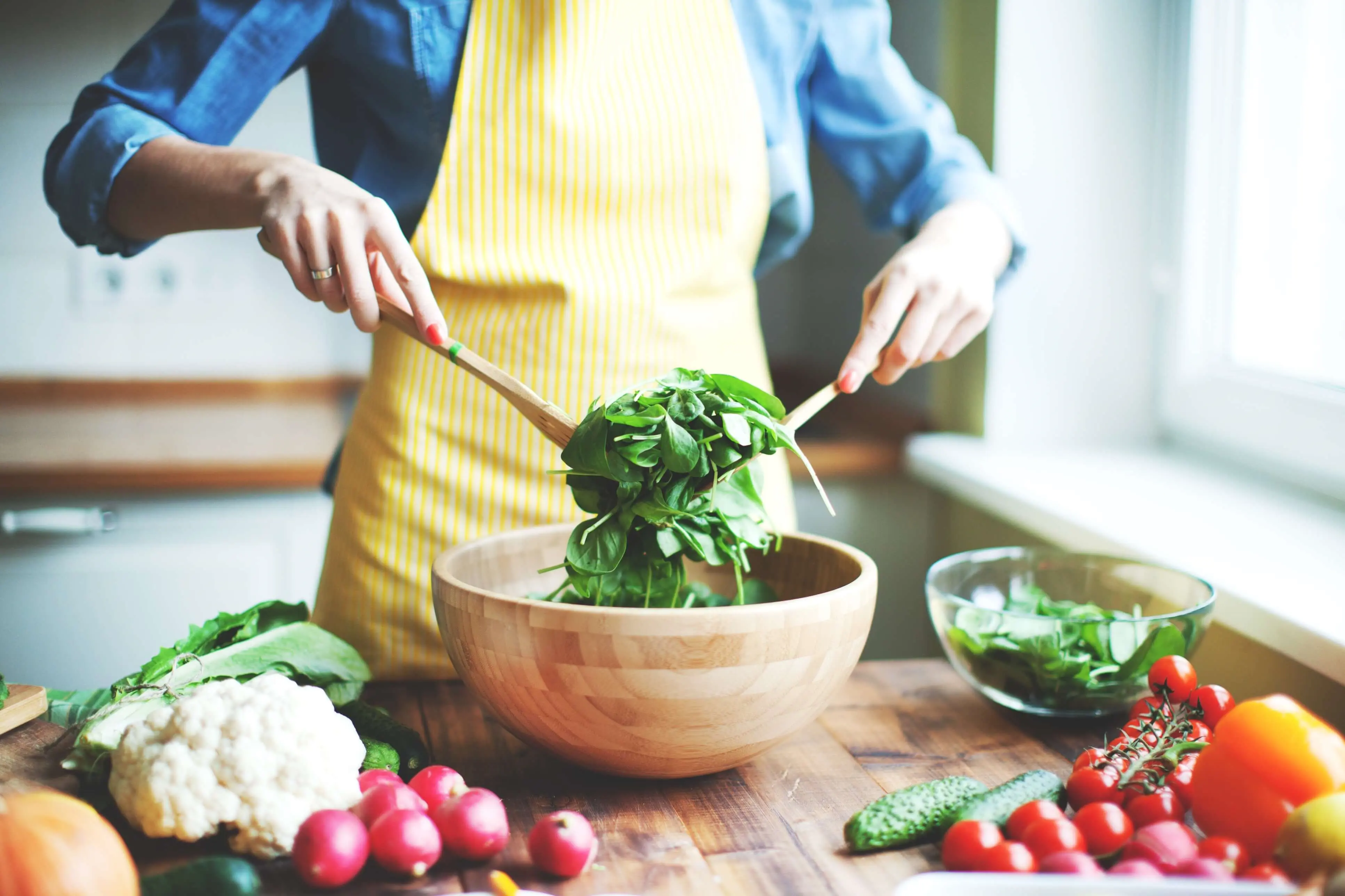Une femme qui porte un tablier jaune remue une salade verte dans un bol en bois. 