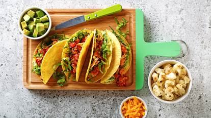 Tacos de chou-fleur affichés sur une planche à découper avec des bols latéraux remplis de fromage, d'avocat et de chou-fleur.