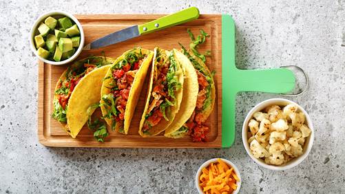 Tacos de chou-fleur affichés sur une planche à découper avec des bols latéraux remplis de fromage, d'avocat et de chou-fleur.