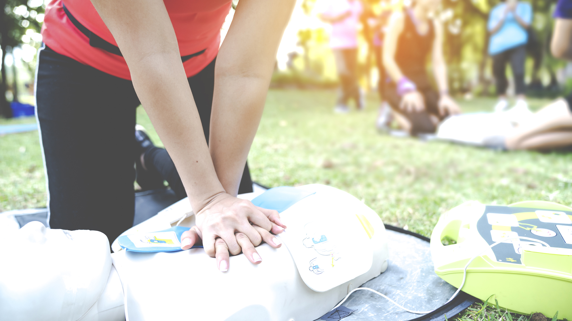 Une femme suit un cours sur la RCR dans un parc; les doigts entrelacés, elle administre des compressions thoraciques à un mannequin sur lequel un DEA est mis en place.
