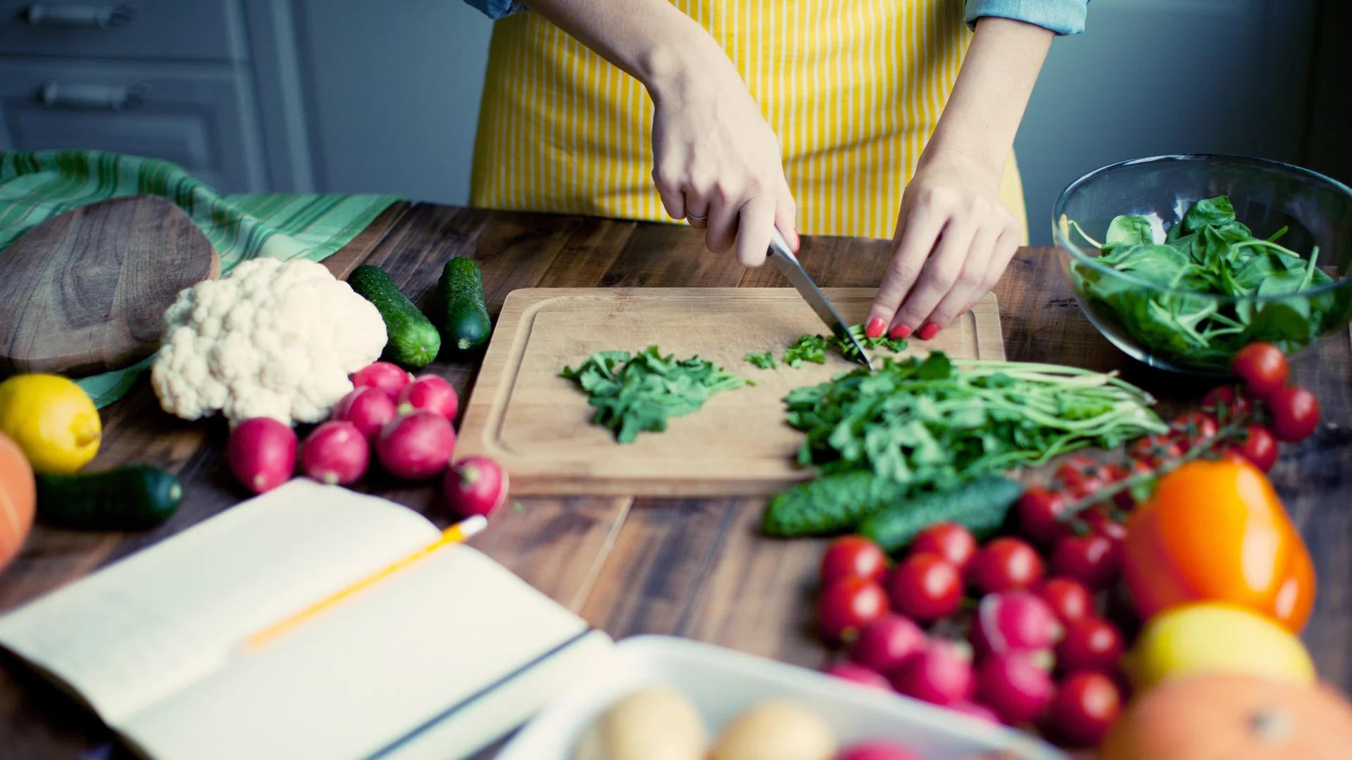 Une personne coupe des légumes sur une planche à découper, avec une grande variété de légumes frais et un livre de recettes ouvert sur la table.