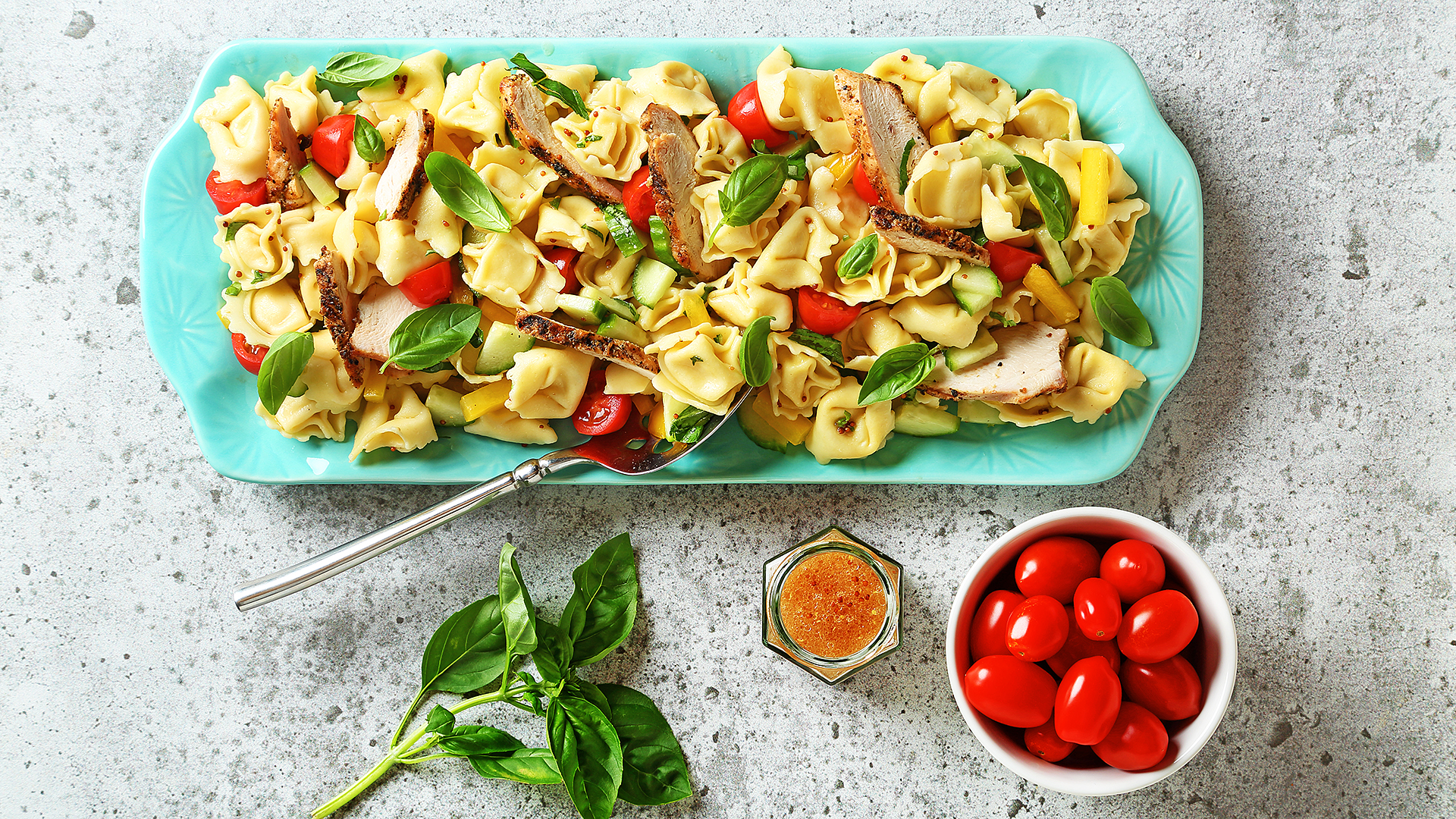 Salade de tortellini à la méditerranéenne sur un plat de service bleu avec un bol de tomates cerises, une vinaigrette et un brin de basilic sur le côté.