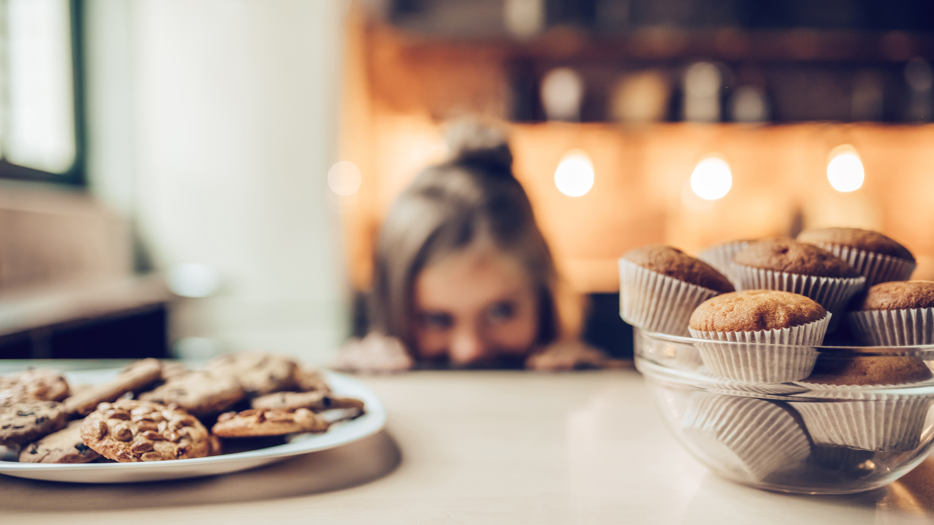 Une assiette de biscuits et un bol de muffins sont fixés du regard par une fillette en arrière-plan.