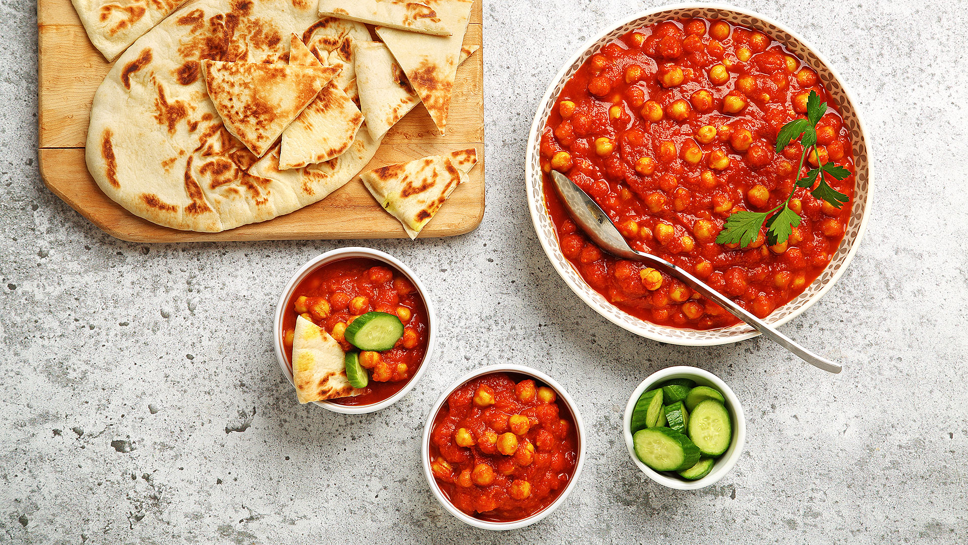 Pois chiches aux tomates et à l’ail dans un grand bol de service avec, sur le côté, du pain naan sur une planche à découper en bois et des tranches de concombre dans un petit bol.