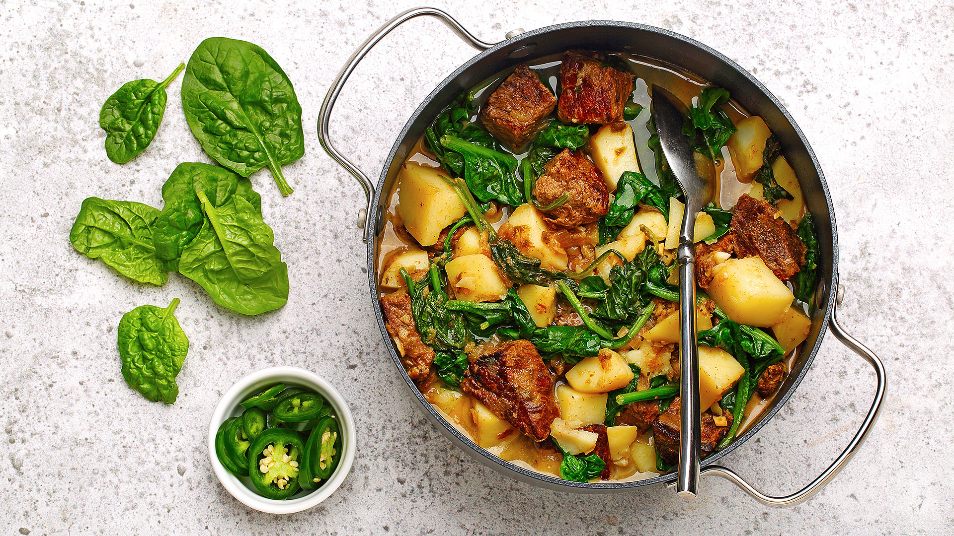 Bœuf épicé avec épinards et pommes de terre dans un poêlon avec des feuilles d’épinards et du jalapeño coupé présenté sur le côté.