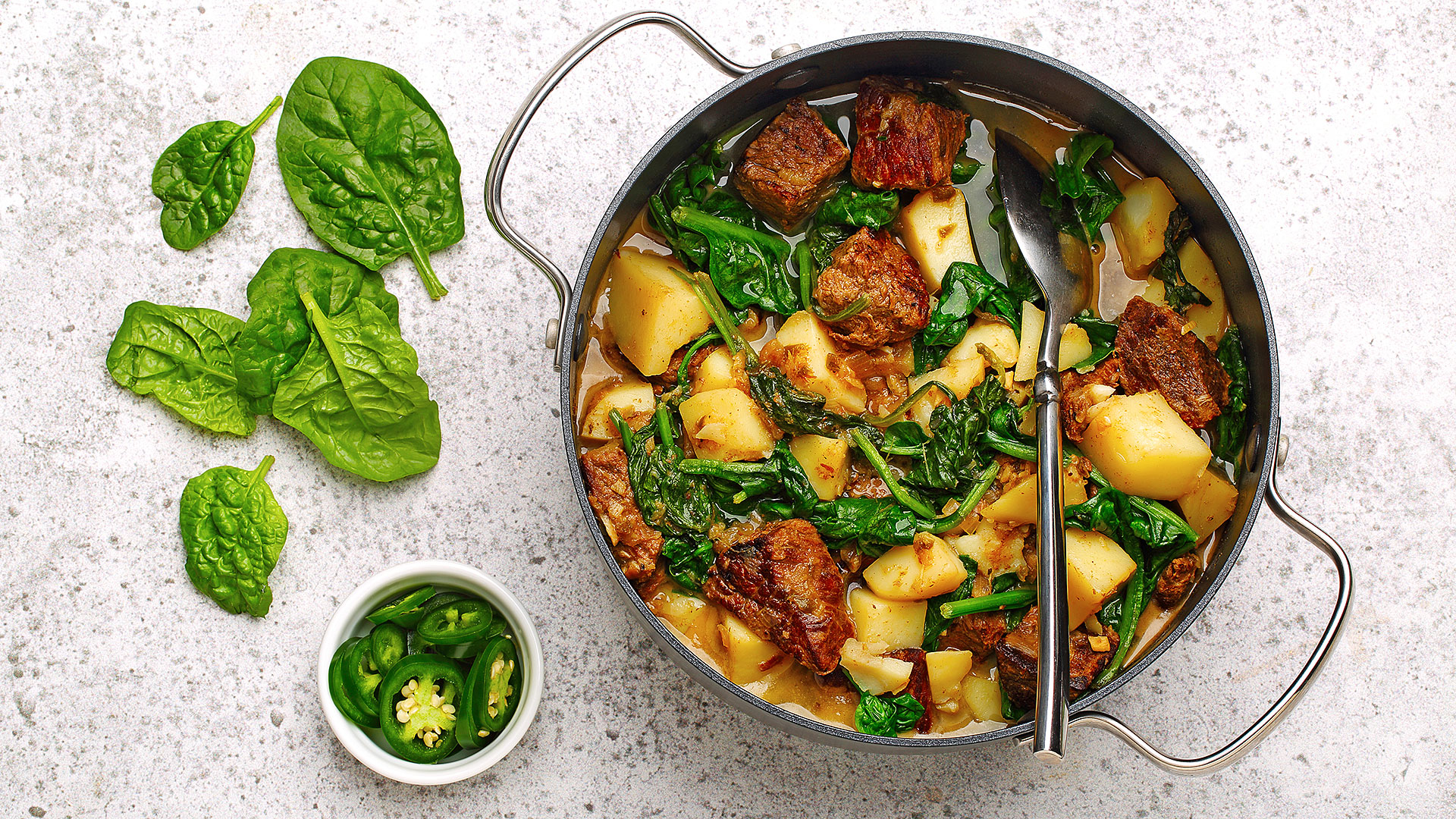 Bœuf épicé avec épinards et pommes de terre dans un poêlon avec des feuilles d’épinards et du jalapeño coupé présenté sur le côté. 