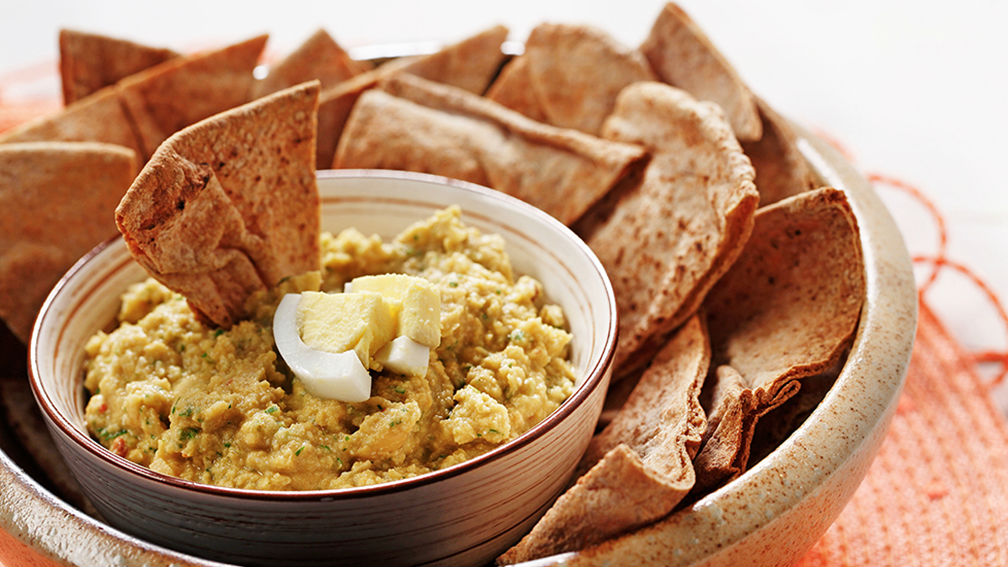 Bol de houmous avec tranches oeuf dur sur le dessus assis à l'intérieur plus grand bol rempli de l'ensemble du pain pita de blé.