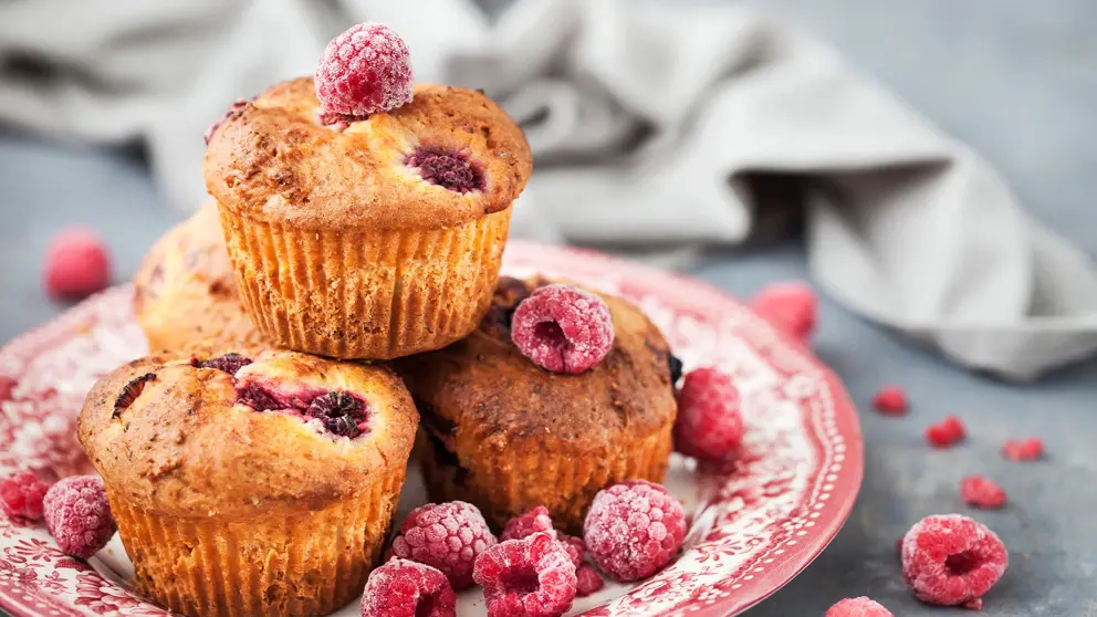 Quatre muffins aux framboises sur un plat garni de framboises congelées. 
