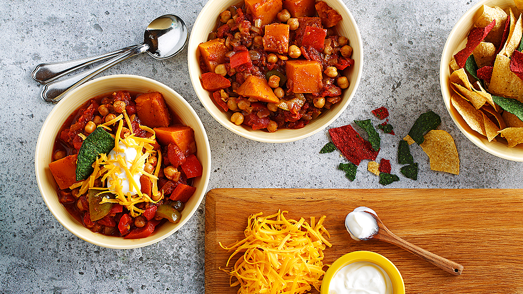 Deux bols de chili reposant sur un comptoir, à côté d’une planche en bois couverte de fromage râpé et de crème sure, et d’un bol rempli de croustilles de maïs.