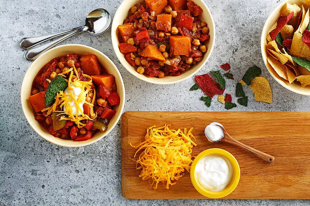 Un plat ovale orange à poignées contenant le ragoût de haricots, de patates douces et d’ail, à côté d’une cuillère en bois et de deux bols pour le service