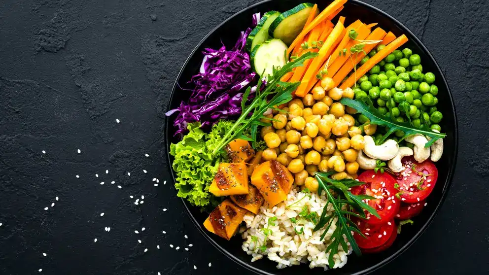 Un grand bol sur fond sombre déborde de légumes dont des pois chiches, des carottes, du chou et des tomates.