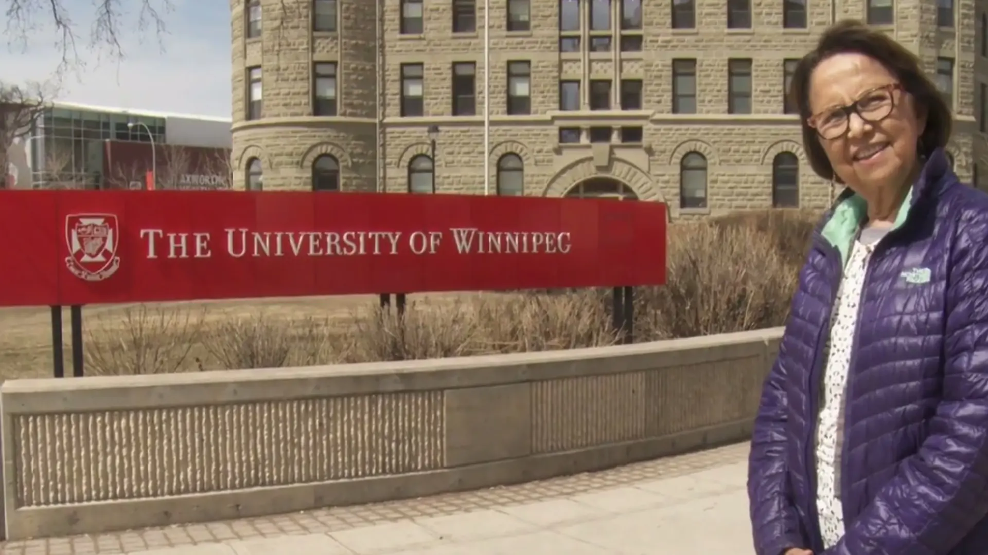 Esther Sanderson sourit, habillée d’une veste violette, à côté d’un panneau de l’Université de Winnipeg. 