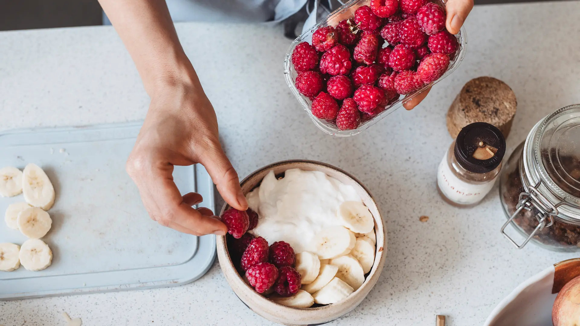 Une personne dépose des fraises dans un bol avec du yogourt et des bananes.