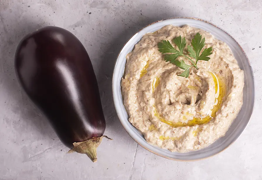 Une aubergine entière à côté d’un bol de trempette à l’aubergine garnie de persil et d’un filet d’huile d’olive.