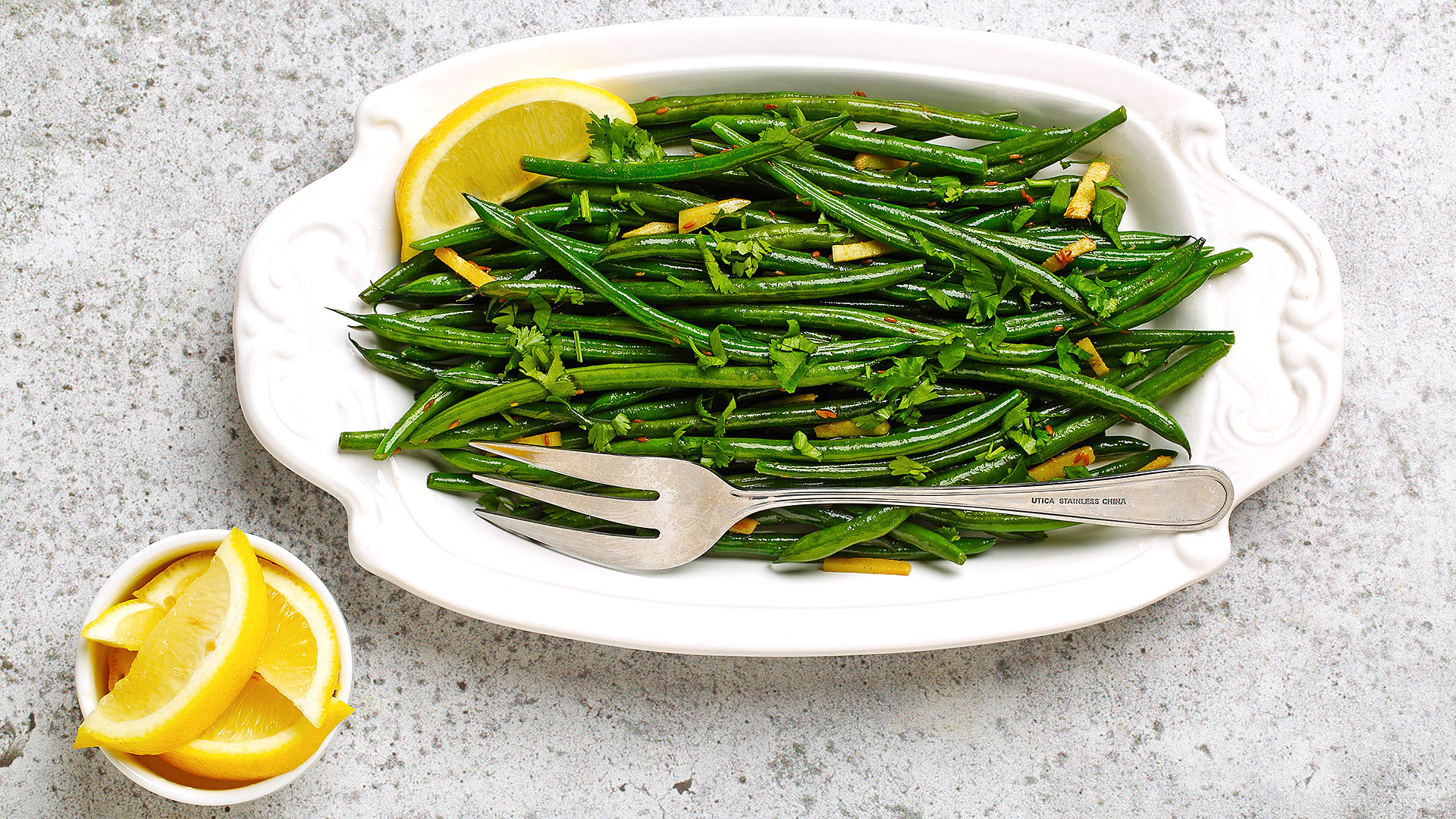 Haricots verts rissolés au gingembre dans un plat de service blanc, avec une fourchette en argent et des tranches de citron sur le côté. 