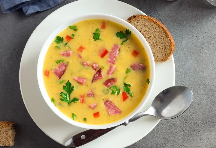 Chaudrée de jambon et de pommes de terre dans un bol blanc accompagnée d’une tranche de pain.