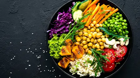 Un grand bol sur fond sombre déborde de légumes dont des pois chiches, des carottes, du chou et des tomates.