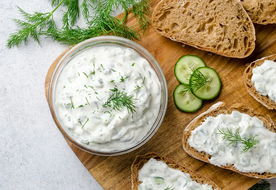Un bol de trempette ranch au yogourt grec garnie d’aneth et des tranches de pain tartinées de trempette sur une planche à découper en bois.