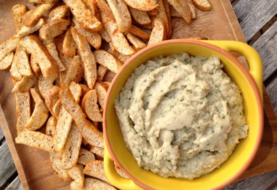 Un bol de houmous de petits haricots ronds blancs à côté d’une pile de croustilles de bagel assaisonnées sur un plateau en bois.
