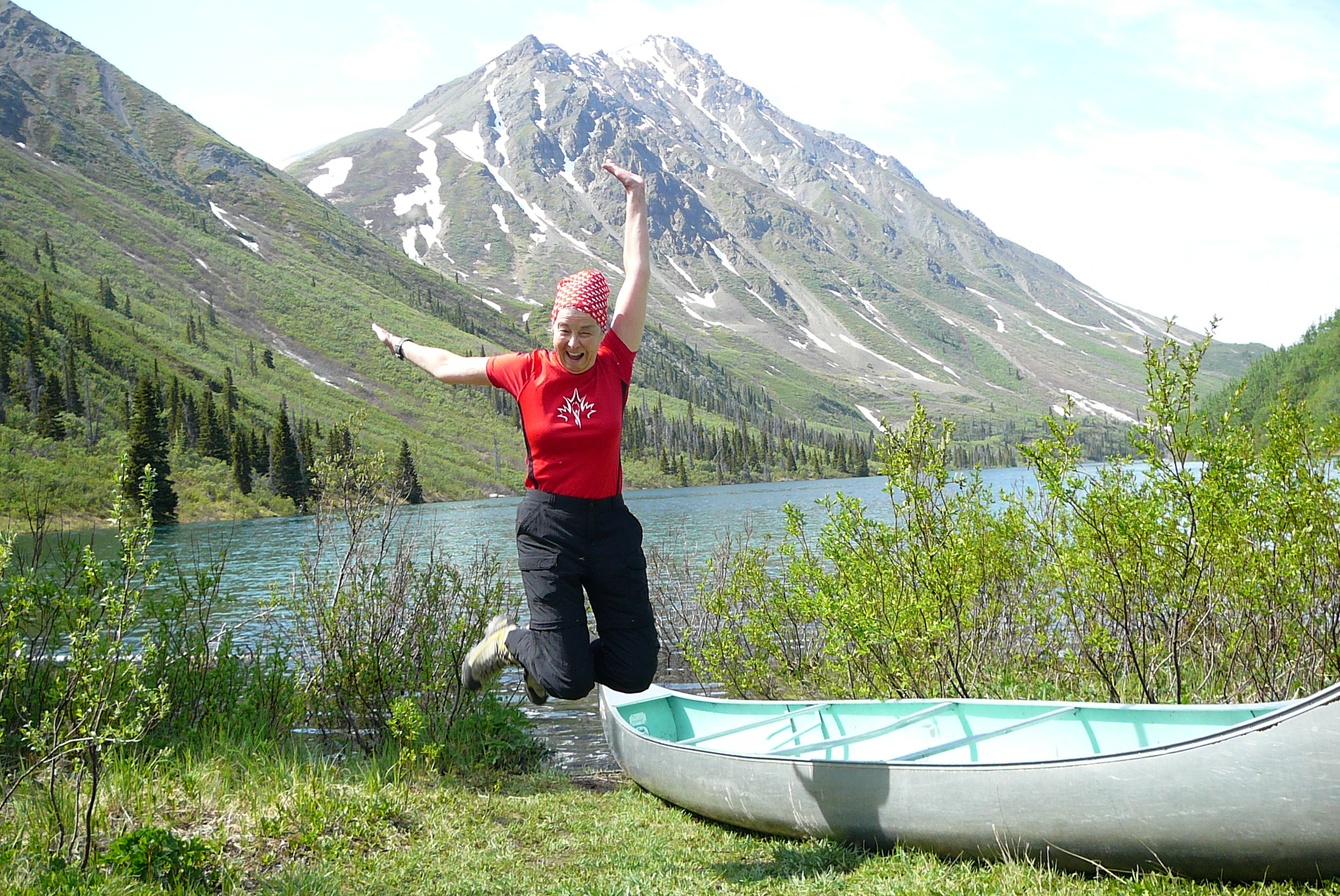 Donna, souriante, saute dans les airs à côté d’un canoë, devant une montagne.