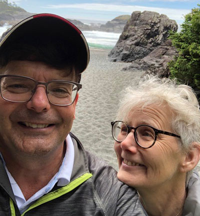 Un homme et une femme âgés sourient côte à côte sur une plage de sable parsemée de rochers.