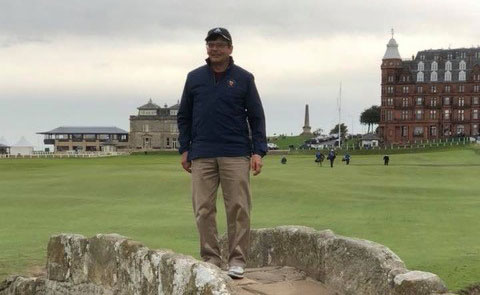 Un homme se tient sur un pont en pierre au milieu d’un terrain de golf sous un ciel gris.
