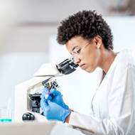 Jeune femme noire dans une blouse de laboratoire regarde à travers un microscop