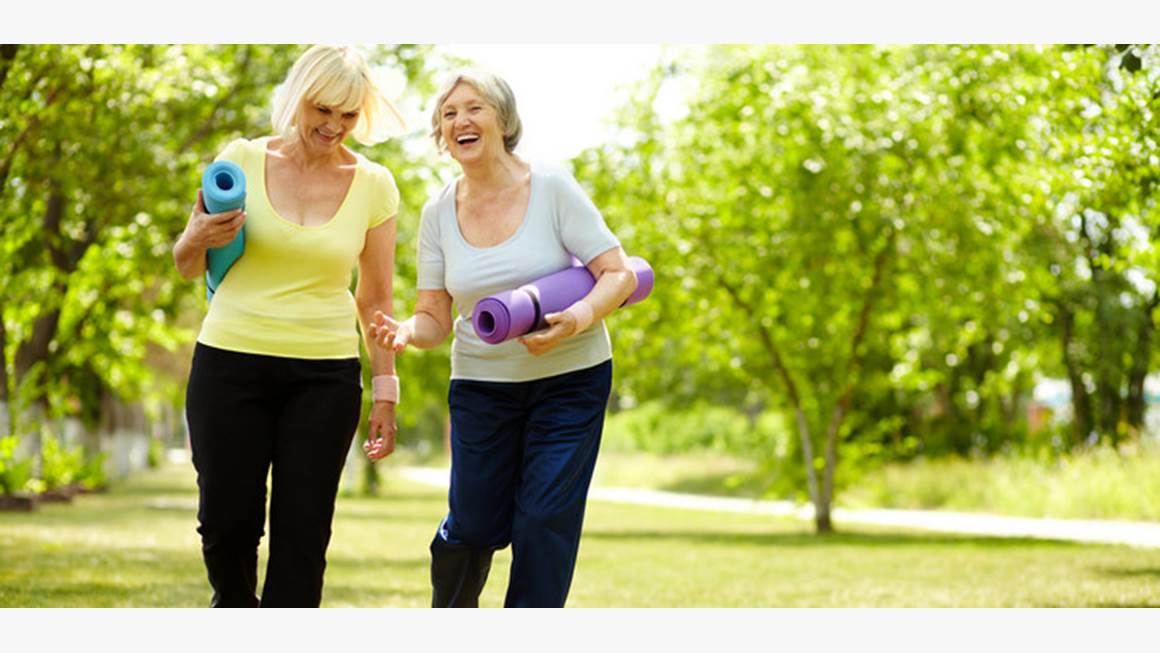 Two older women carrying yoga mats stroll through a park in the summertime, laughing. 